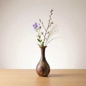 May include: A brown ceramic vase holds a delicate floral arrangement. The arrangement includes a single lavender-colored flower, sprigs, and blades of grass. The vase has a narrow neck and a rounded body, sitting on a wooden surface.