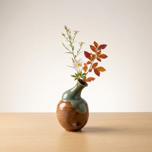 Puede incluir: Un jarrón de cerámica hecho a mano con un cuello curvo único. El jarrón presenta un diseño de dos tonos, con una base marrón y una sección superior verde. Contiene un delicado arreglo de flores blancas y hojas rojas y naranjas vibrantes.