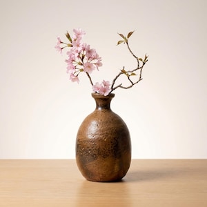 May include: A brown, textured ceramic vase with a narrow neck, holding branches with delicate pink cherry blossoms and green leaves. The vase sits on a light-colored wooden surface, creating a natural, minimalist aesthetic.