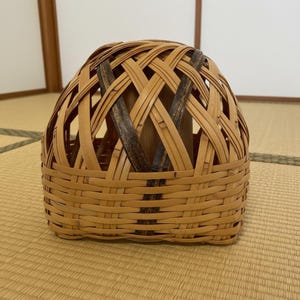 May include: A hand-woven bamboo basket with a rounded top and a square base. The basket has a lattice design with alternating light and dark brown bamboo strips. The basket is on a light brown woven mat.