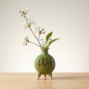 Puede incluir: Un jarrón de cerámica verde con cuerpo redondo y tres patas cortas. El jarrón contiene una rama de flores blancas, pequeñas flores azules y hojas verdes. El jarrón tiene un diseño texturizado en marrón.