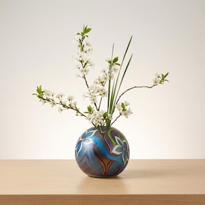 Puede incluir: Un jarrón redondo y decorativo con un diseño floral azul, verde y marrón. El jarrón contiene ramas de flores blancas y hojas verdes. El jarrón está sobre una superficie de madera marrón claro sobre un fondo blanco.