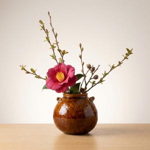 May include: A brown ceramic vase with a glossy finish holds a vibrant arrangement. A large, deep pink flower with a yellow center is the focal point, complemented by slender branches with green leaves and buds. The vase sits on a wooden surface.