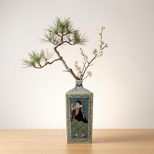 May include: A rectangular ceramic vase with a blue and white patterned rim and a painted scene of a figure in traditional clothing. The vase holds a branch of white flowers and a pine branch.