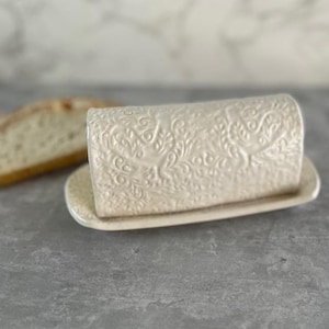 Puede incluir: Un mantequillero de cerámica blanca con un patrón floral decorativo. El mantequillero tiene una tapa y una base.