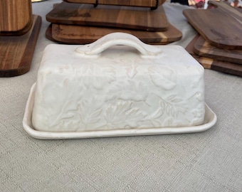 May include: A white ceramic butter dish with a lid and handle, featuring a raised leaf pattern. The dish sits on a matching rectangular base. Wooden cutting boards are visible in the background.