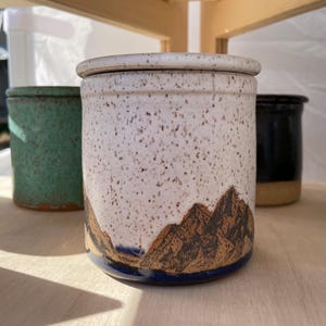 May include: A cylindrical ceramic canister with a lid, featuring a speckled off-white upper section and a dark brown mountain range illustration at the base. The bottom edge shows a thin band of deep blue glaze, contrasting with the earthy tones.