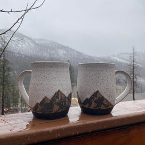 Puede incluir: Dos tazas de cerámica blanca con diseños de cordilleras negras y esmalte blanco moteado. Las tazas están sobre una superficie de madera con una cordillera nevada en el fondo.