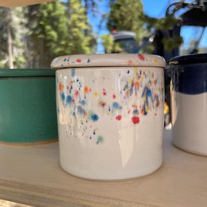 May include: A cylindrical ceramic jar with a matching lid, featuring a glossy off-white base glaze decorated with a colourful splatter pattern in shades of blue, orange, yellow, and red. The container has a smooth, straight-sided exterior.