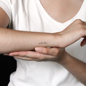 May include: A small, black, cursive temporary tattoo on the inner forearm featuring the word aloha in a delicate, lowercase script font. The design is simple and minimalist, positioned horizontally on the skin against a plain white background.