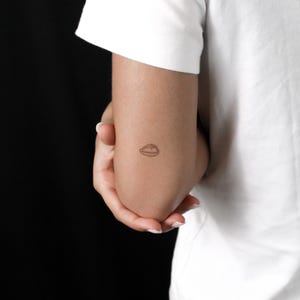 Puede incluir: Un pequeño tatuaje temporal de tinta negra con un dibujo lineal sencillo de una concha marina. El tatuaje está situado en la parte superior del brazo y muestra una estética de líneas finas y limpias con sutiles detalles de puntos.