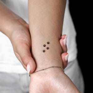 May include: A temporary tattoo featuring a small, black, minimalist design of three animal paw prints arranged in a diagonal line. The tattoo is applied to the skin of a wrist, showcasing a clean, simple graphic element with fine lines.