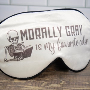 K&ouml;nnte beinhalten: Wei&szlig;e Schlafmaske mit einem schwarzen Skelett, das ein Buch liest, und dem Text "Morally Gray is my favorite color".