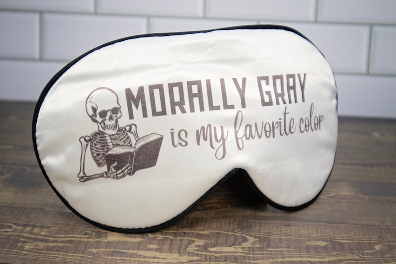 K&ouml;nnte beinhalten: Wei&szlig;e Schlafmaske mit einem schwarzen Skelett, das ein Buch liest, und dem Text "Morally Gray is my favorite color".