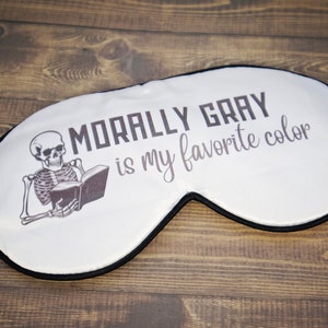 K&ouml;nnte beinhalten: Wei&szlig;e Schlafmaske mit einem schwarzen Skelett, das ein Buch liest, und dem Text "Morally Gray is my favorite color".