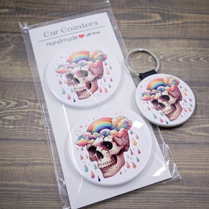 Puede incluir: Conjunto de dos posavasos redondos para coche con un cráneo con un arcoíris y nubes en la parte superior. Los posavasos son blancos con un diseño colorido y el texto "Car Coasters Handmade with love" está impreso en el embalaje.