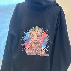 May include: A navy blue hooded sweatshirt featuring a colorful graphic of a cartoon character. The character is brown with green hair and is sitting in front of a splash of blue, red, and pink paint.