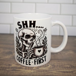 May include: White ceramic coffee mug with a black and white illustration of a skeleton holding a coffee cup with a heart on it. The text "SHH... COFFEE FIRST" is printed on the mug.
