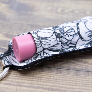 Könnte beinhalten: Ein Stoff-Lippenbalsamhalter mit einem schwarz-weißen Blumenmuster, befestigt an einem silbernen Schlüsselring. Ein rosa Lippenbalsam ist teilweise sichtbar. Der Halter liegt auf einer Holzoberfläche.