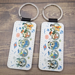 Op de afbeelding: Twee rechthoekige sleutelhangers met een witte achtergrond en een nautisch thema met Mickey Mouse, Donald Duck en Pluto. Elke sleutelhanger heeft een zilveren ring en een zwarte band.