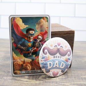 May include: A decorative card with a superhero skeleton illustration and the text "THE HERO" next to a round button with a mustache design and the text "BEST DAD". The items are on a wooden surface.