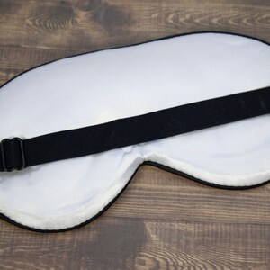 K&ouml;nnte beinhalten: Wei&szlig;e Schlafmaske mit einem verstellbaren schwarzen Band. Die Maske besteht aus weichem, bequemem Material und ist so konzipiert, dass sie Licht abh&auml;lt.