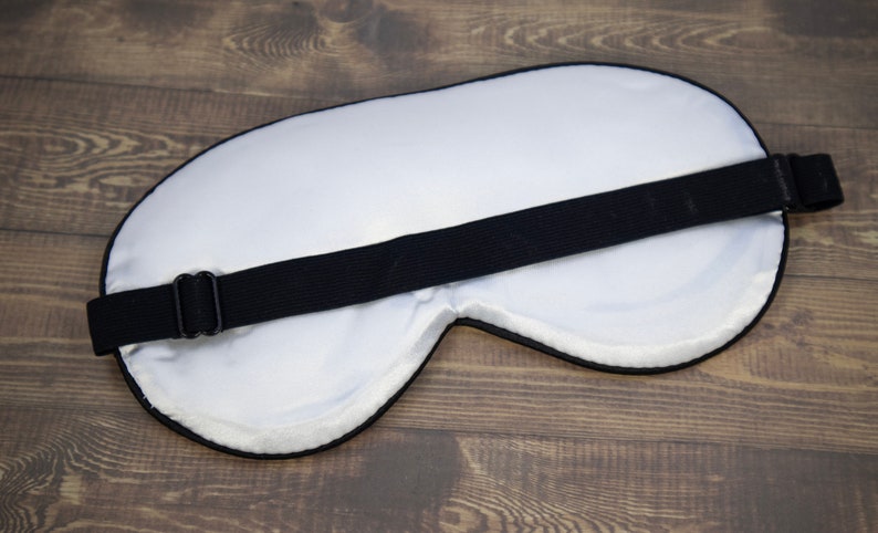 K&ouml;nnte beinhalten: Wei&szlig;e Schlafmaske mit einem verstellbaren schwarzen Band. Die Maske besteht aus weichem, bequemem Material und ist so konzipiert, dass sie Licht abh&auml;lt.