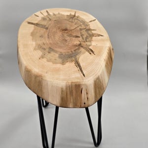 May include: A side table featuring a natural live-edge wood slab top with visible grain patterns and dark radial cracks. The tabletop is supported by three slender, black metal hairpin legs that provide a stable base for the circular wood piece.