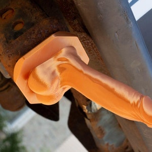 May include: An orange, phallic-shaped object, possibly a novelty item or a decorative piece, is attached to a rusty metal structure. The object's smooth surface contrasts with the rough, aged metal.