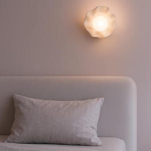 Può includere: Una lampada da parete bianca a forma di fiore emette una luce soffusa in una camera da letto. Sotto, un cuscino grigio chiaro poggia su un letto con lenzuola abbinate. L'atmosfera è minimalista e serena.