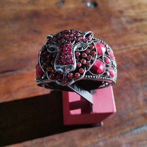 May include: A silver and red jeweled bracelet with a tiger head design. The bracelet is displayed on a red stand.
