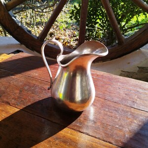 Peut inclure: Un pichet en argent avec un corps arrondi et une anse incurvée posé sur une table en bois. Le pichet reflète la lumière, créant un effet brillant.