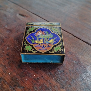 Könnte beinhalten: Eine kleine, rechteckige Schachtel mit einem blauen, schwarzen und goldenen Emaille-Design. Das Design zeigt ein stilisiertes Blumenmotiv mit Vögeln und Wellen. Die Schachtel hat einen Klappdeckel und ein blaues Innenleben.