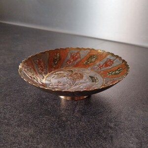 Puede incluir: Un pequeño cuenco decorativo de latón con un borde festoneado. El cuenco está decorado con un patrón floral en tonos naranja, verde y blanco.