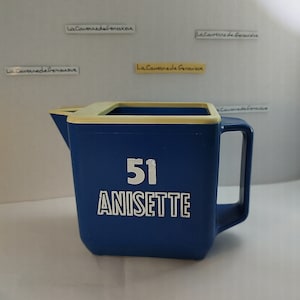 Puede incluir: Jarra azul con borde y asa color crema. La jarra tiene el número "51" y la palabra "ANISETTE" impresa en blanco en la parte delantera. La jarra probablemente se utiliza para servir bebidas.
