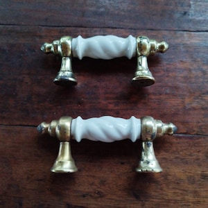 May include: Two white ceramic drawer pulls with a twisted design and gold metal accents. The pulls are on a wooden surface.