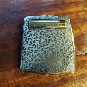 Puede incluir: Un encendedor de cigarrillos de plata con un diseño dorado ornamentado. El encendedor tiene una forma rectangular y una tapa con bisagras.