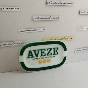 Könnte beinhalten: Ein weißer Aschenbecher aus Keramik mit grünem Rand und einem grünen Band um die Mitte. Der Aschenbecher trägt die Aufschrift "AVEZE AUVERGNE GENTIANE" in schwarzen Buchstaben. Auf dem Aschenbecher sind drei gelbe Blumen abgebildet.
