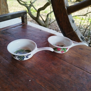 May include: Two white ceramic bowls with handles. One bowl has a black and green berry design, the other has a red and green raspberry design.