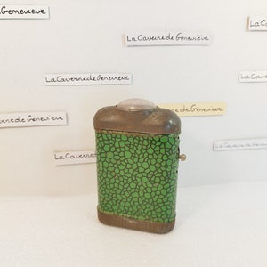 Könnte beinhalten: Vintage Metallbehälter mit grünem und schwarzem Muster und einem transparenten, abgerundeten Deckel. Der rechteckige Behälter hat einen Klappdeckel und einen kleinen Metallknopf. Im Hintergrund sind kleine Papieretiketten mit dem Text "La Caverne de Genevieve" zu sehen.