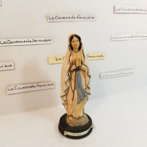 May include: A white figurine of the Virgin Mary, standing on a black base with the text "NOTRE-DAME DE LOURDES" in gold lettering. The figurine is wearing a blue and white robe and holding a rosary.