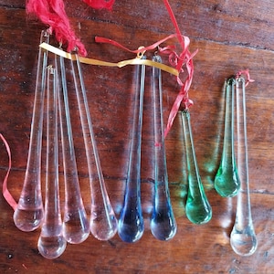Peut inclure: Une collection de huit ornements en verre en forme de larme de différentes couleurs, dont transparent, rose, bleu et vert. Les ornements sont liés ensemble avec un ruban rouge et or.