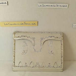 Pode incluir: Uma caixa de cartão branca com guarnições douradas e um design decorativo. A caixa tem o texto "La Caverne de Genevieve" impresso nela.