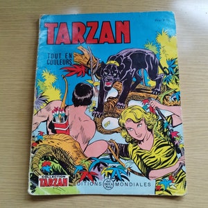 以下が含まれることがあります： ターザン、女性、そして黒いパンサーを描いた、ビンテージなコミックの表紙。表紙は青色で、タイトルの「TARZAN」は大きな白い文字で書かれている。「TOUT EN COULEURS」という文字がタイトルの下にある。このコミックは「COLLECTION TARZAN」シリーズのもので、「EDITIONS DUCA MONDIALES」によって出版されている。