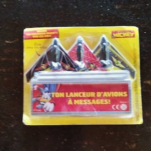 May include: A yellow and red package with a Mickey Mouse character on it. The package contains a toy airplane launcher with the text "Ton Lanceur D'Avions A Messages!" on the front.