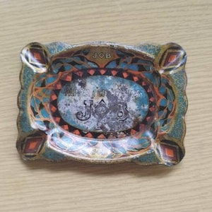 Op de afbeelding: Een vintage metalen asbak met een kleurrijk ontwerp met de tekst "JOB" en "PAPIERS A CIGARETES" in een cirkelvormig frame. De asbak heeft een blauwe, oranje en gouden kleurstelling.