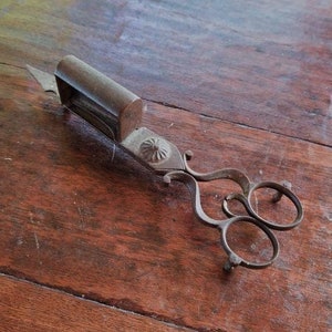 May include: Antique metal fireplace tool with a long handle and a pair of tongs at the end. The tongs have two loops and a decorative design.