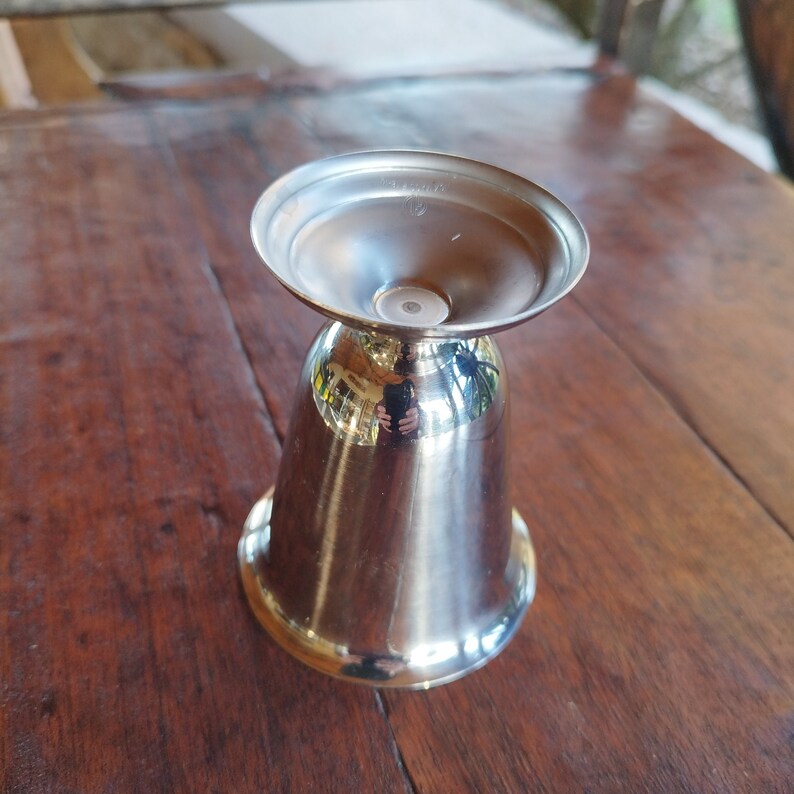 May include: A silver metal jigger with a flared top and a rounded bottom. The jigger is sitting on a wooden surface.