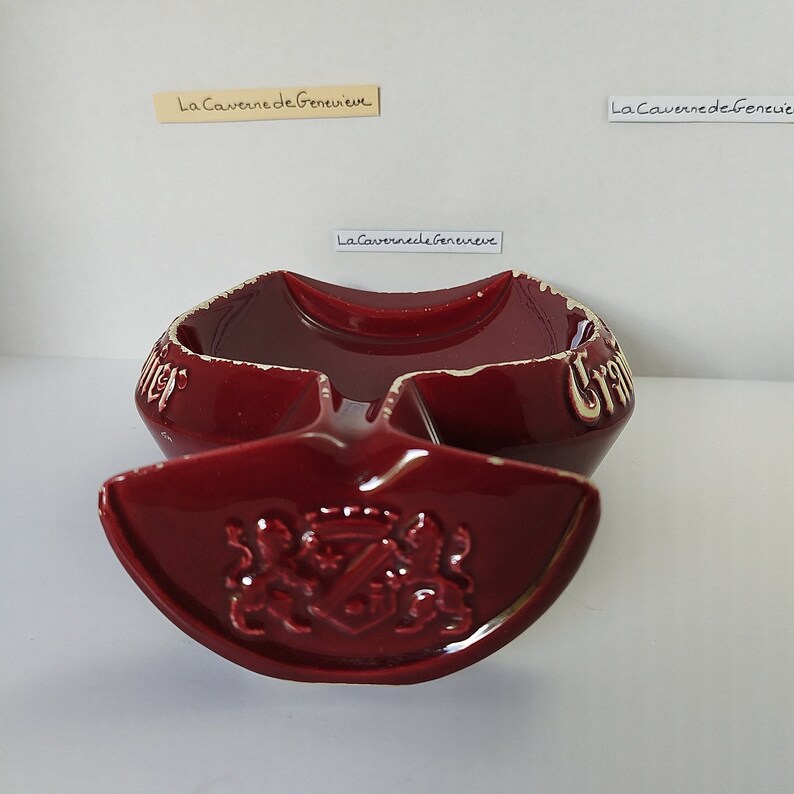 May include: A red ceramic ashtray with a white crest and the text "La Caverne de Geneve" in white. The ashtray has a unique shape with a small bowl and a larger, curved section.