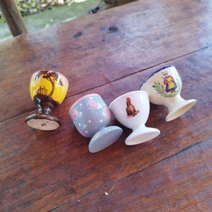 Può includere: Quattro porta uova in ceramica con diversi disegni. Uno è giallo con una silhouette marrone di una casa e alberi, uno è blu con pois bianchi e rose rosa, uno è bianco con un gallo e uno è bianco con un disegno blu e bianco di una donna in un vestito.